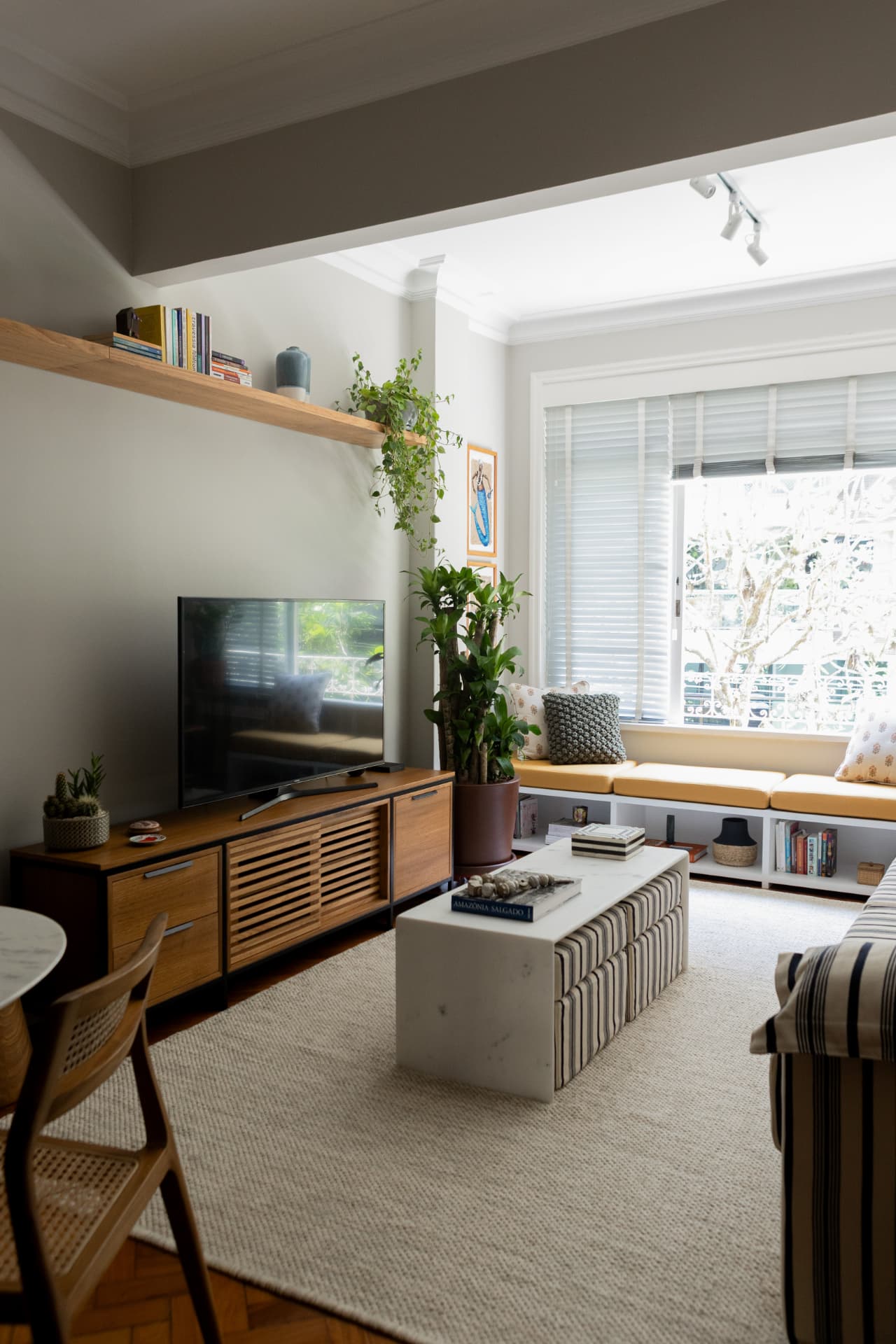 Sala de televisão com banco junto à janela, plantas e mobiliário em madeira